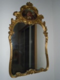 Mirror with an oil painting at the top in a gold frame.