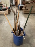Plastic garbage can full of vintage yard tools; wood handle rake, shovel, pic axe and more.