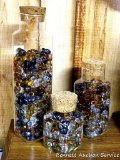 Three glass jars of decorative marbles. Each jar has a cork top. Tallest jar is 13