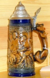 Covered beer stein made in West Germany shows a wild boar hunting scene and has a fox handle. Bottom