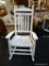 (BACK) WHITE WOODEN ROCKING CHAIR; TALL BACK WITH CURVED TOP RAIL, WIDE ARMS, AND SLATTED SEAT. THE