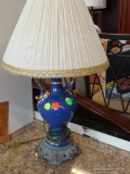 BLUE TABLE LAMP WITH GOLD TONE DETAILING; CREAM COLORED PLEATED, BELL SHAPED SHADE SITTING ATOP A