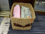BASKET FULL OF ASSORTED LINENS; THIS RECTANGULAR BASKET HAS A HANDLE ON EACH SIDE. IT IS FILLED WITH