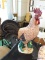LARGE DECORATIVE ROOSTER; MAUVE-TAN IN COLOR WITH RED COMB, BLACK TAIL FEATHERS, AND GREEN BASE.