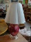 CRANBERRY GLASS TABLE LAMP; WHITE PLEATED LAMP SHADE WITH SCALLOPED BOTTOM EDGE SITTING ATOP A LARGE