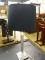METAL TABLE LAMP; BLACK LINEN BELL SHAPED SHADE SITTING ON A BRUSHED SILVER TONED COLUMN STYLE BODY