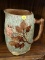 MAJOLICA PITCHER; HAS CHOCOLATE GLAZED AND WINE COLORED FLOWERS WITH A BRANCH STYLE HANDLE.