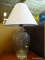 GLASS TABLE LAMP; WHITE PLEATED BELL SHAPED SHADE SITTING ON A LARGE GLASS BODY WITH ROUND METAL