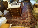 (LR) HAND WOVEN RUG; RED RUG WITH COLORFUL FLORAL PATTERNING AROUND THE RUG AND WHITE TASSEL EDGES