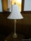 (DEN) BRASS TONE TABLE LAMP; WHITE BELL SHAPED SHADE SITTING ON A TALL CANDLESTICK STYLE BODY WITH