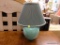 (R2) LAMP; TEAL GLAZED LAMP WITH AN EMERALD AND GOLD TONE COOLIE LAMP SHADE. MEASURES 17 IN TALL.