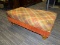 (R3) PLAID OTTOMAN. PLAID AND SUEDE RED AND BROWN OTTOMAN WITH BUTTONS ON SIDES. SITTING ON BUN