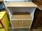 (R2) SIDE TABLE; SIDE TABLE WITH WOOD GRAIN TOP AND WHITE SIDES AND BACK. HAS CUBBY ATOP TWO DOORS