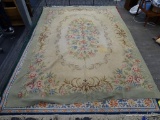 WOVEN AREA RUG; HAS A BEIGE AND GREEN COLOR BASE WITH PINK AND BLUE FLOWERS. HAS BROWN COLORED