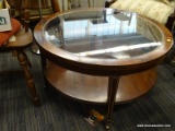 ROUND MAHOGANY COFFEE TABLE WITH GLASS TOP AND 1 LOWER SHELF. IS ON CASTERS. MEASURES 33.5 IN X 18