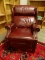 (OFF) BURGUNDY LEATHER UPHOLSTERED RECLINING ARMCHAIR WITH BRASS STUDDING ALONG THE EDGES. MEASURES