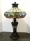 Slagged And Stained Glass Lamp With Bronze Toned Urn Style Base. Primary Colors Include Cream,
