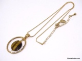 LIA SOPHIA GOLD TONE OVAL PENDANT WITH AN OVAL CUT TIGERS EYE GEMSTONE CENTER, DISPLAYED ON A GOLD